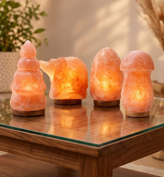 Four glowing salt lamp sculptures in different organic shapes arranged on a glass-topped wooden table in a warmly lit interior setting.